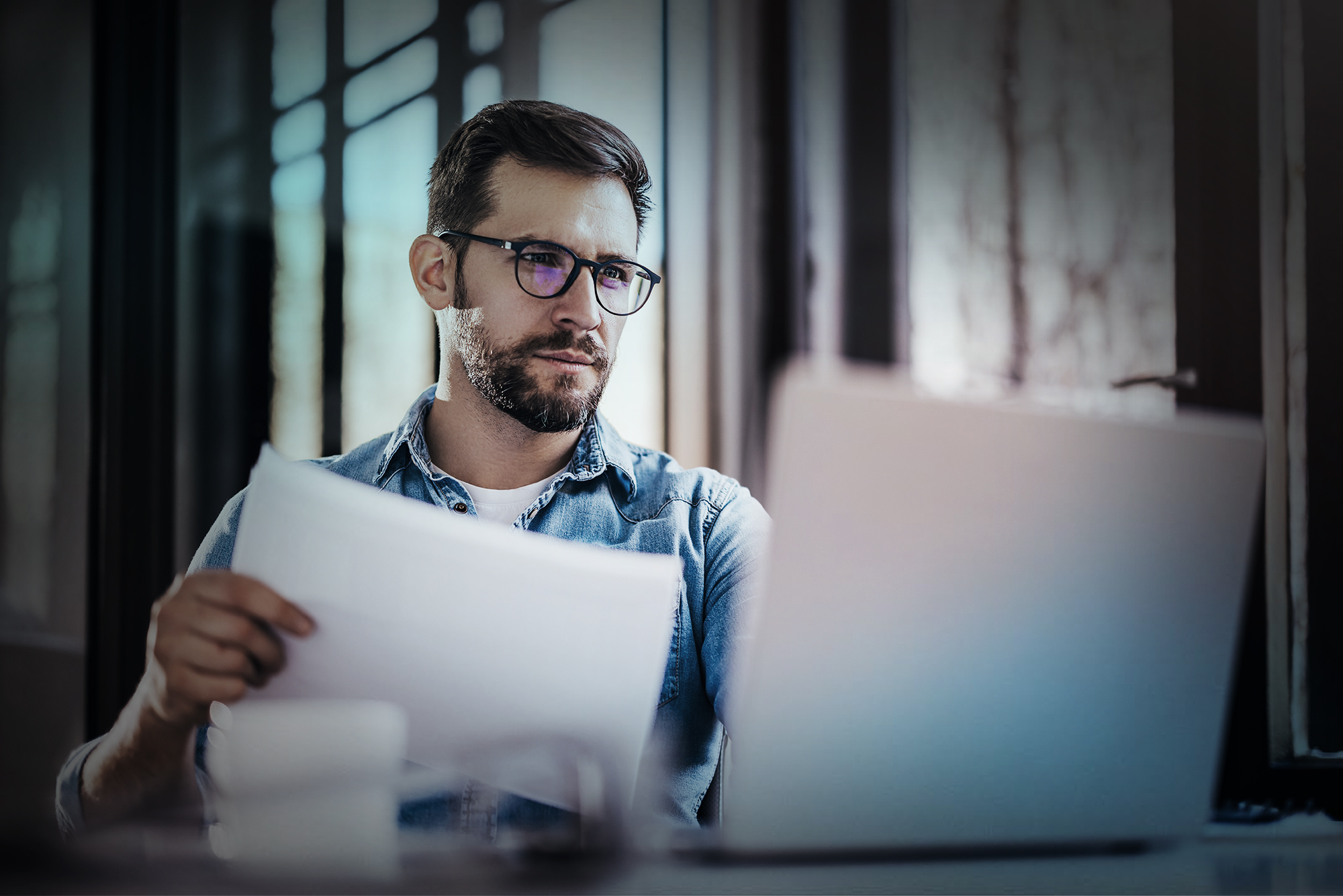 Satisfied young man with glasses sitting at a desk and doing paperwork at his workplace.
